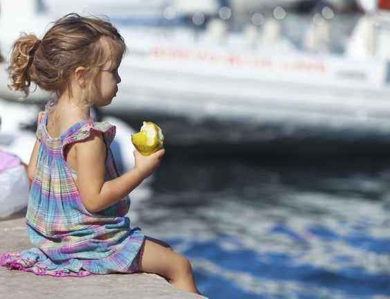 Little blond girl sits on the dock with pear in her hand.